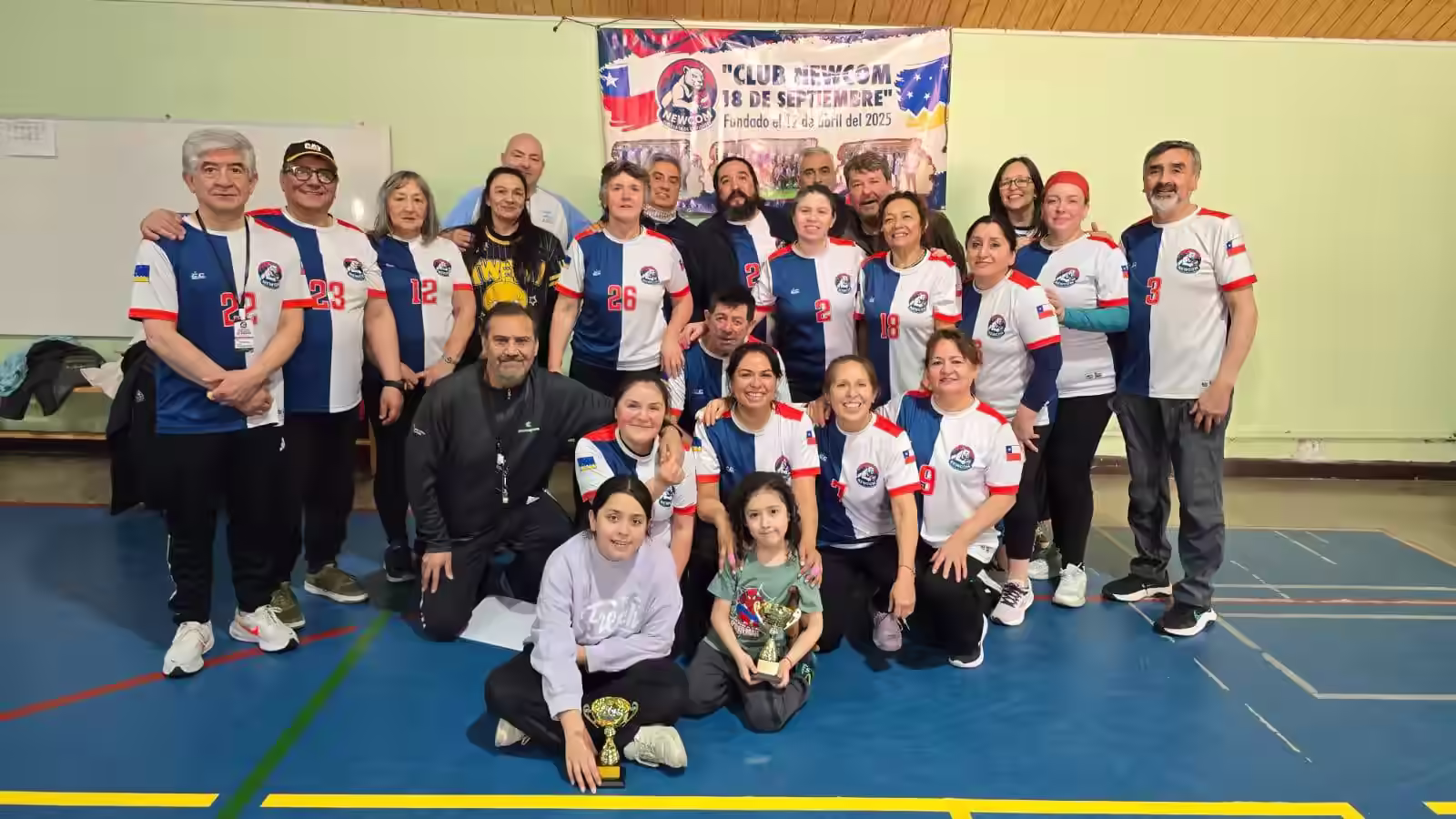 Foto grupal del Club Newcom de Punta Arenas con la bandera del club y los trofeos en el gimnasio
